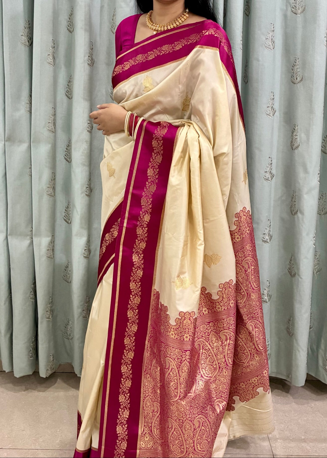 Woman wearing a cream and red saree standing in front of patterned curtains.