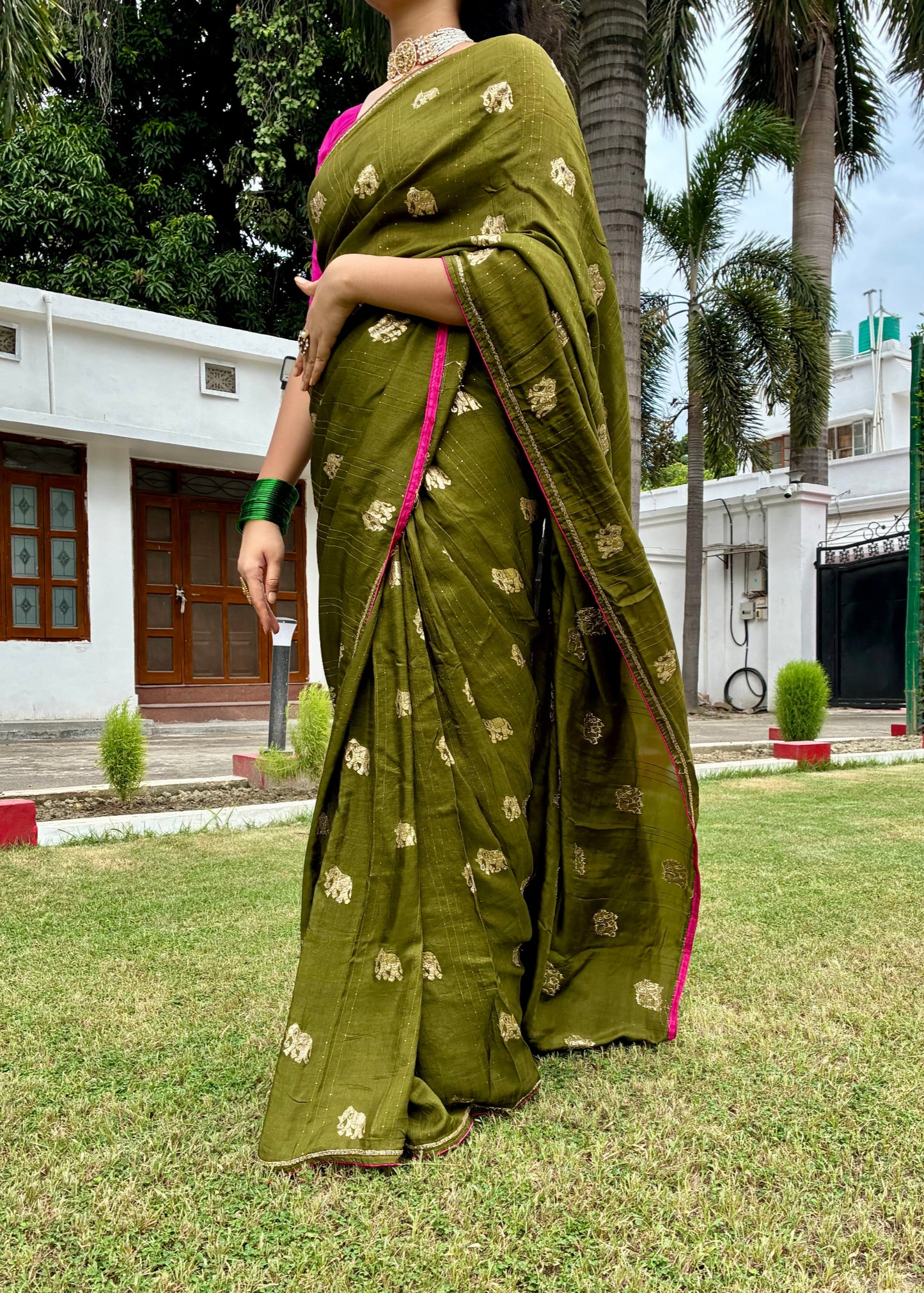 Woman in a green saree with gold patterns standing on grass with trees and buildings in the background
