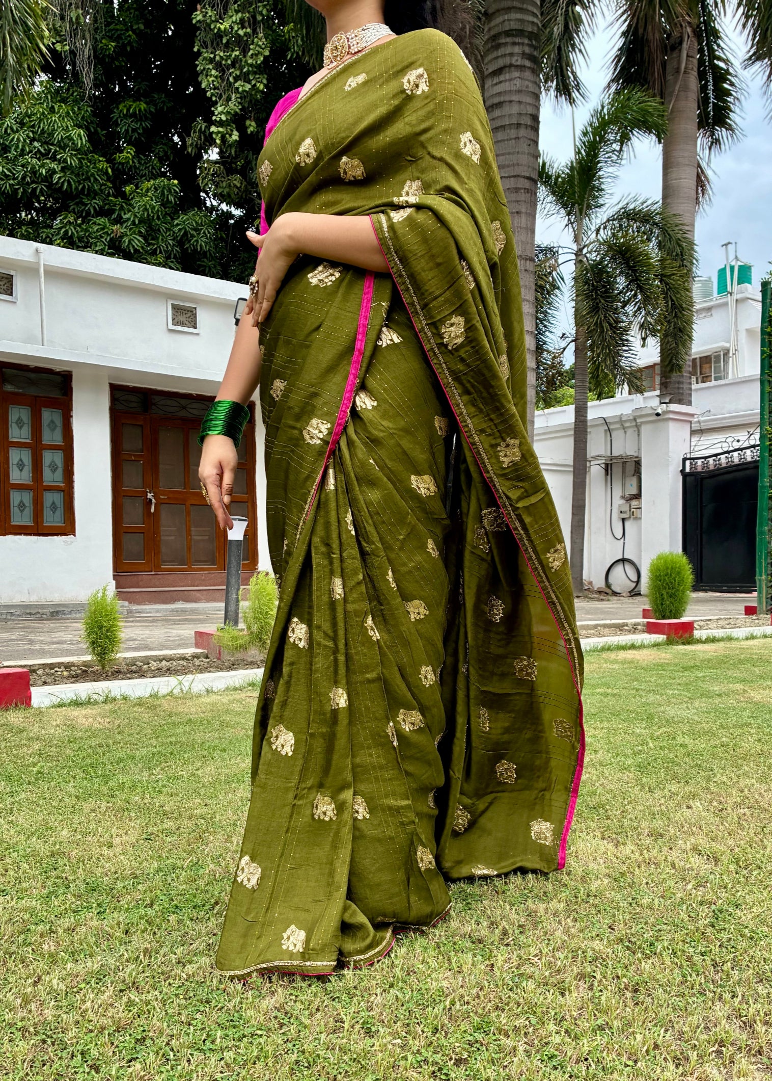 Woman in a green saree with gold patterns standing on grass with trees and buildings in the background