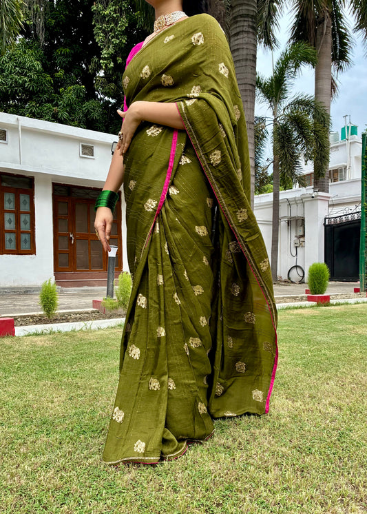 Woman in a green saree with gold patterns standing on grass with trees and buildings in the background