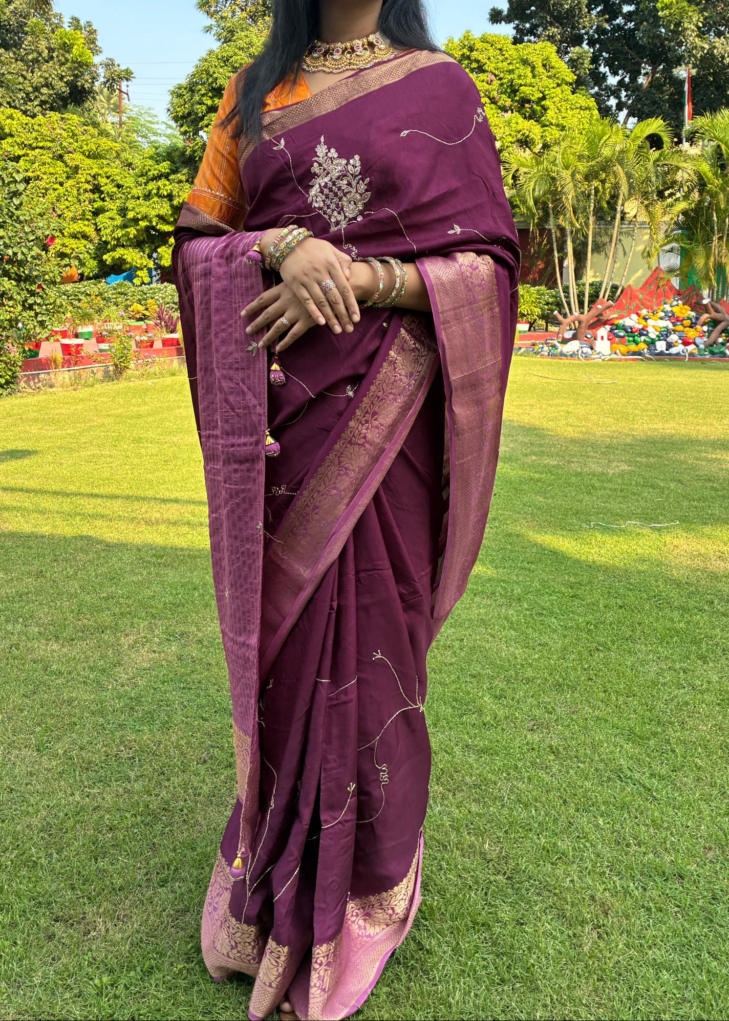 Woman in a purple saree standing outdoors with greenery in the background