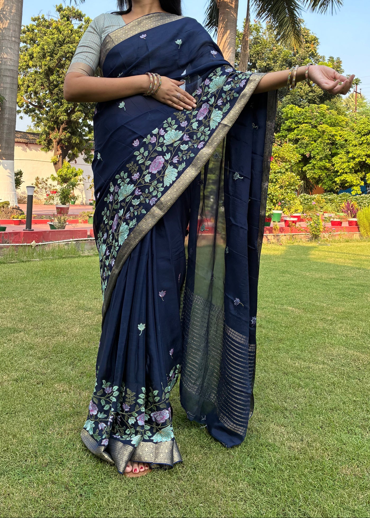 Person holding a dark blue saree with floral patterns in an outdoor setting with green grass and trees.