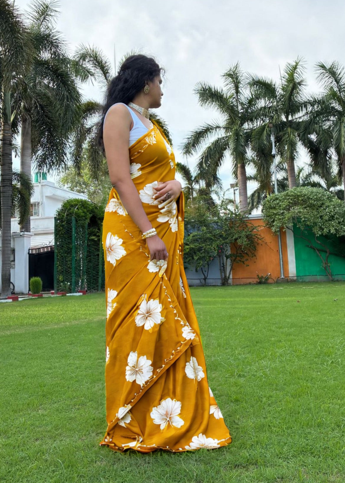 Woman in a yellow saree with white floral patterns standing on grass with palm trees in the background