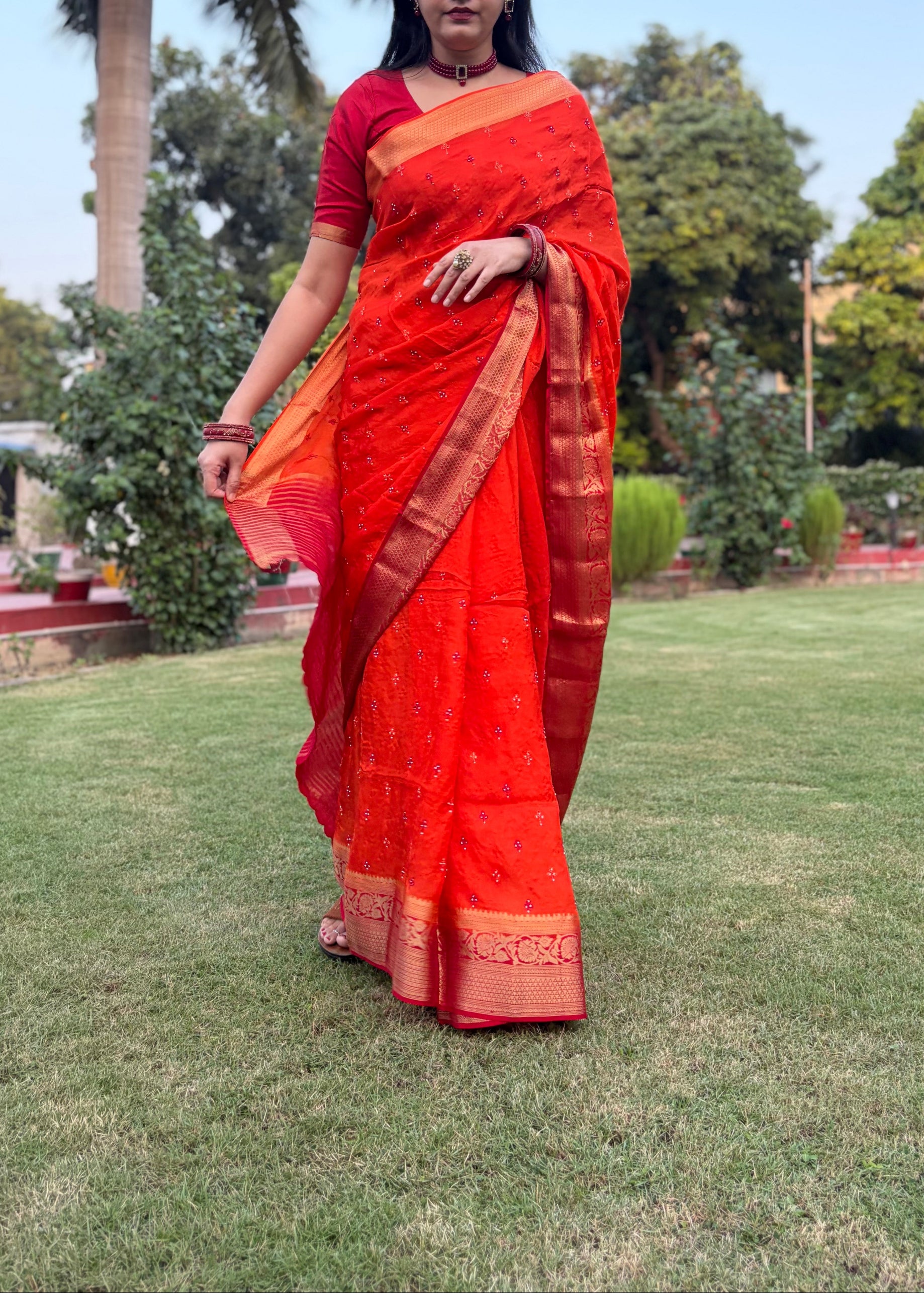 Woman in a bright orange saree standing on grass with trees in the background
