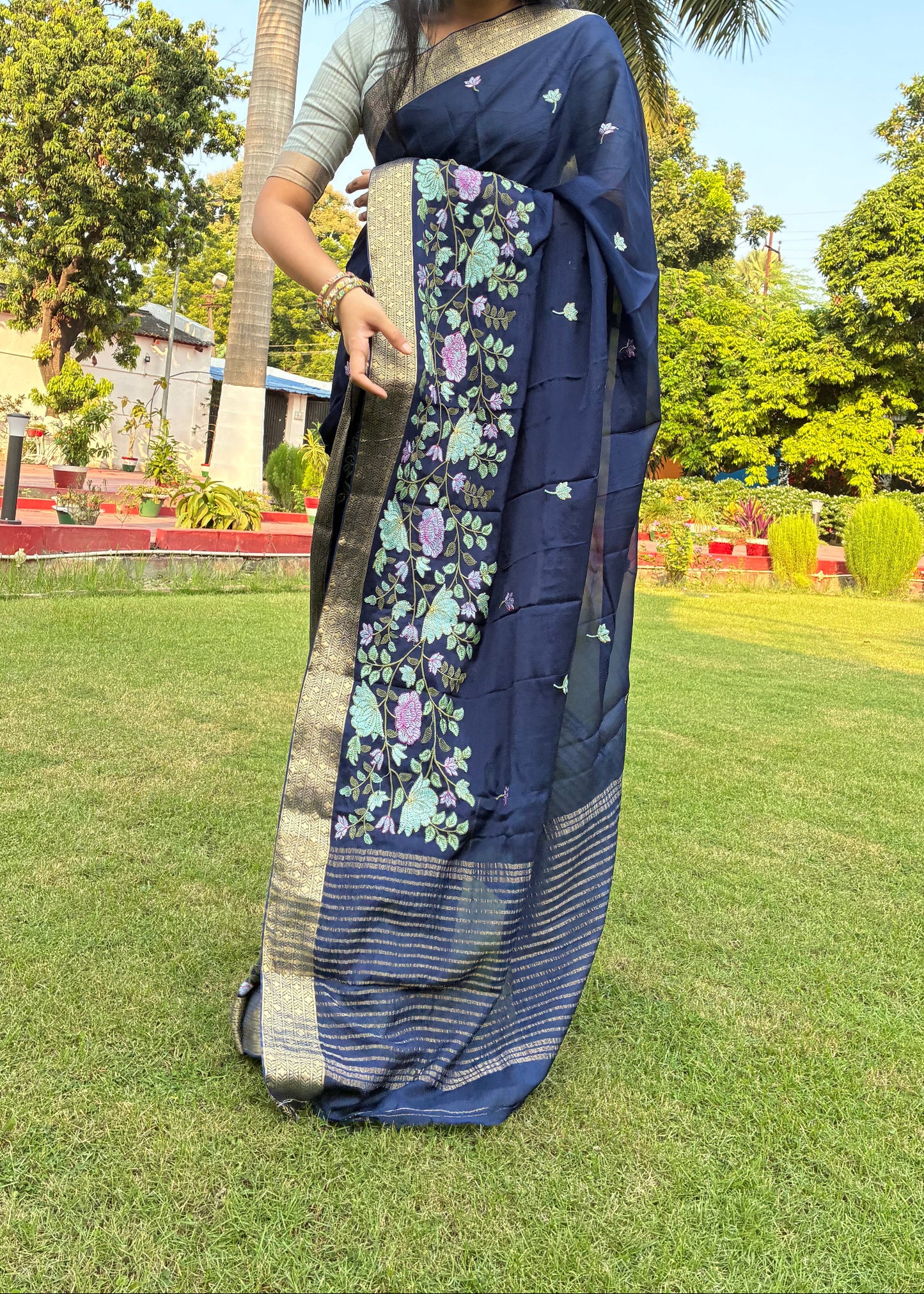 Woman holding a blue saree with floral patterns on grass and trees in the background