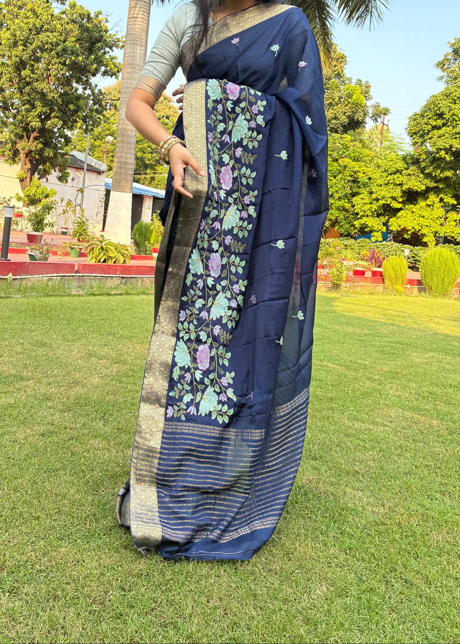 Woman holding a blue saree with floral patterns on grass and trees in the background