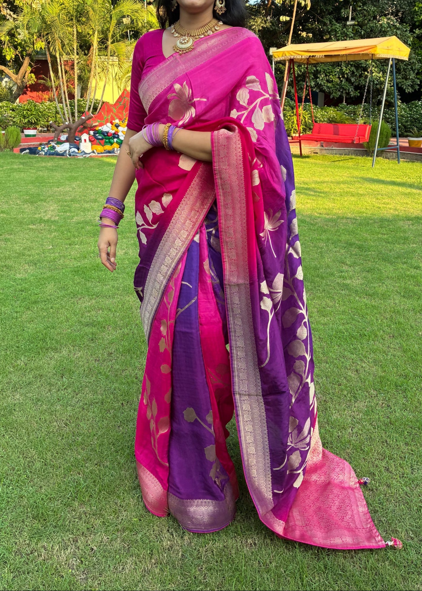Woman in a pink and purple saree standing on grass with trees in the background
