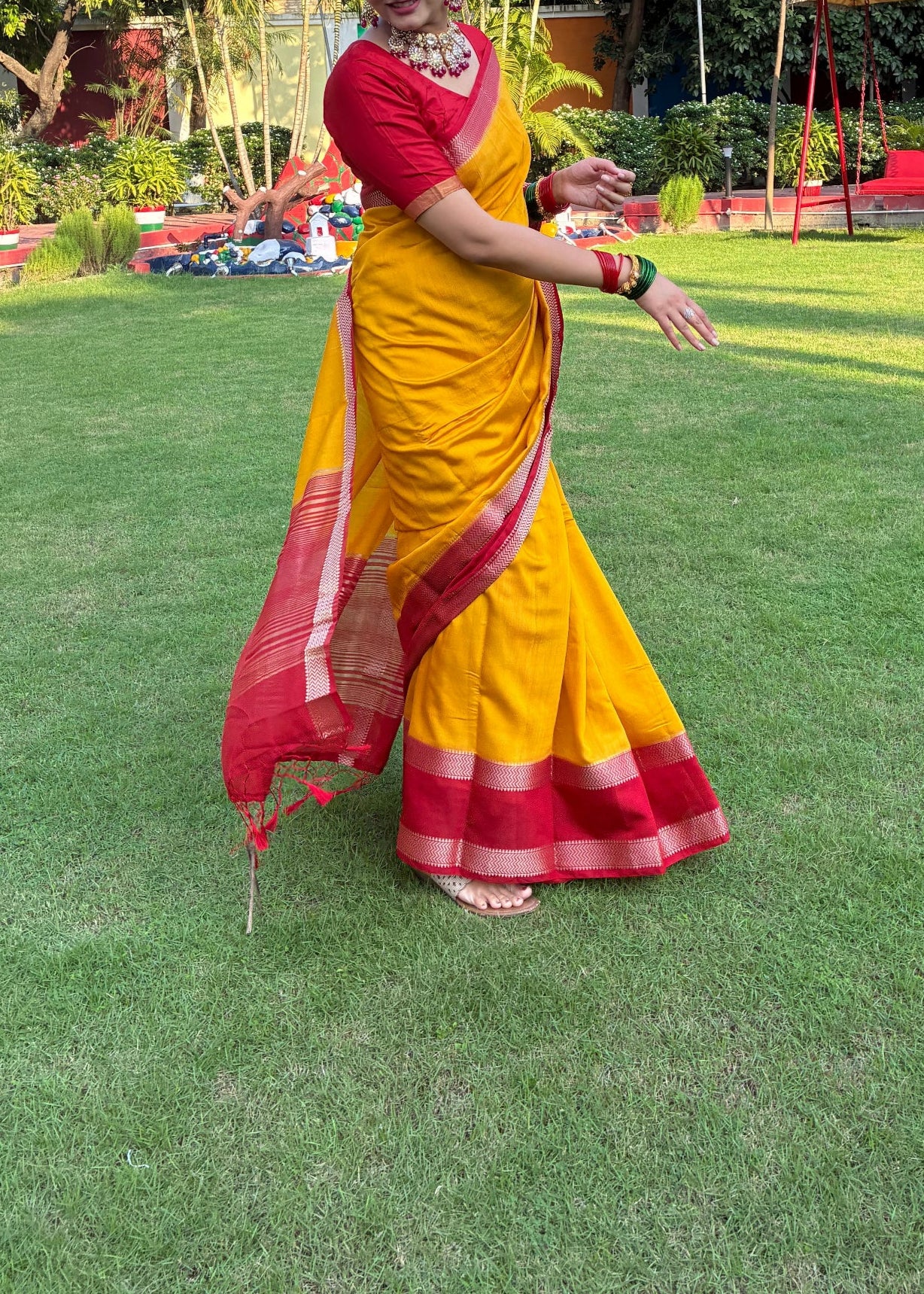 Woman in a yellow and red saree standing on grass with trees in the background