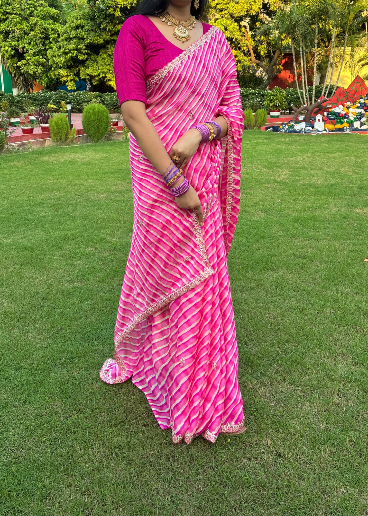 Woman in a pink saree standing on a grassy field with trees and people in the background