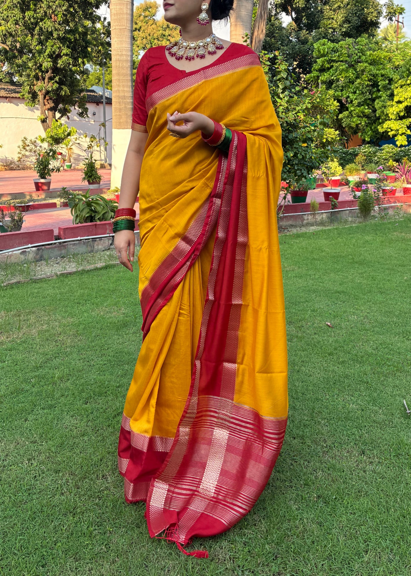 Person wearing a yellow and red saree standing on grass with trees and buildings in the background