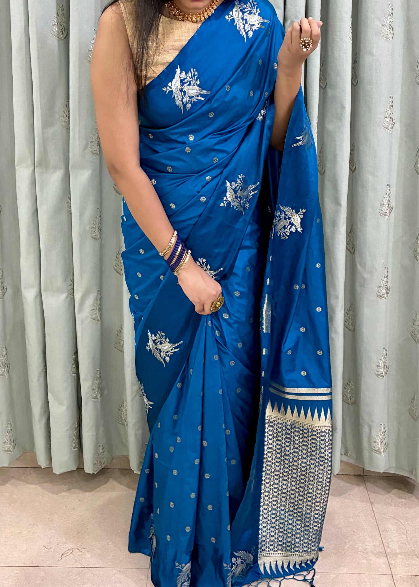 Woman wearing a blue saree with white patterns in front of a light gray curtain.