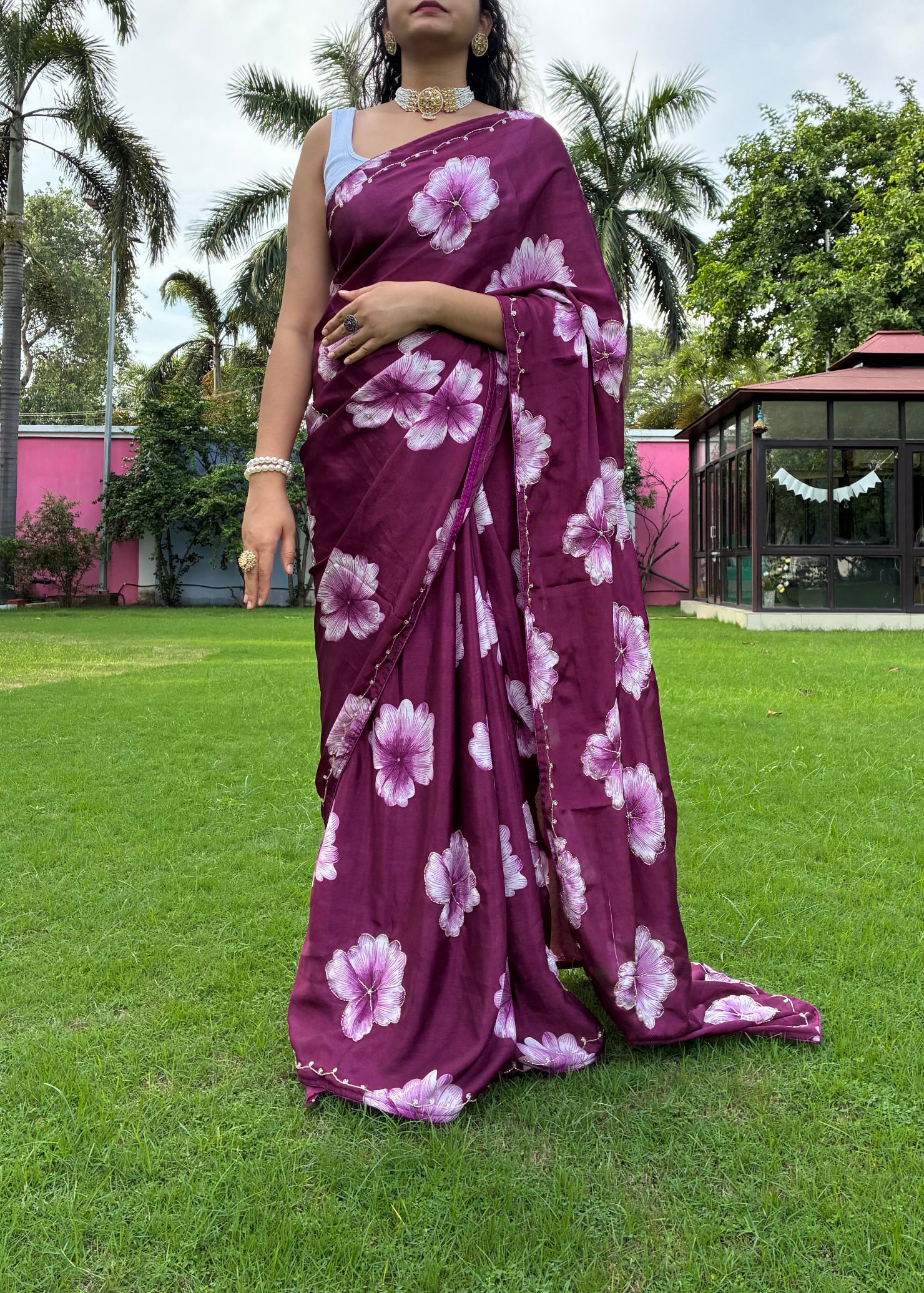 Woman wearing a purple floral saree on a grassy lawn with trees and a building in the background