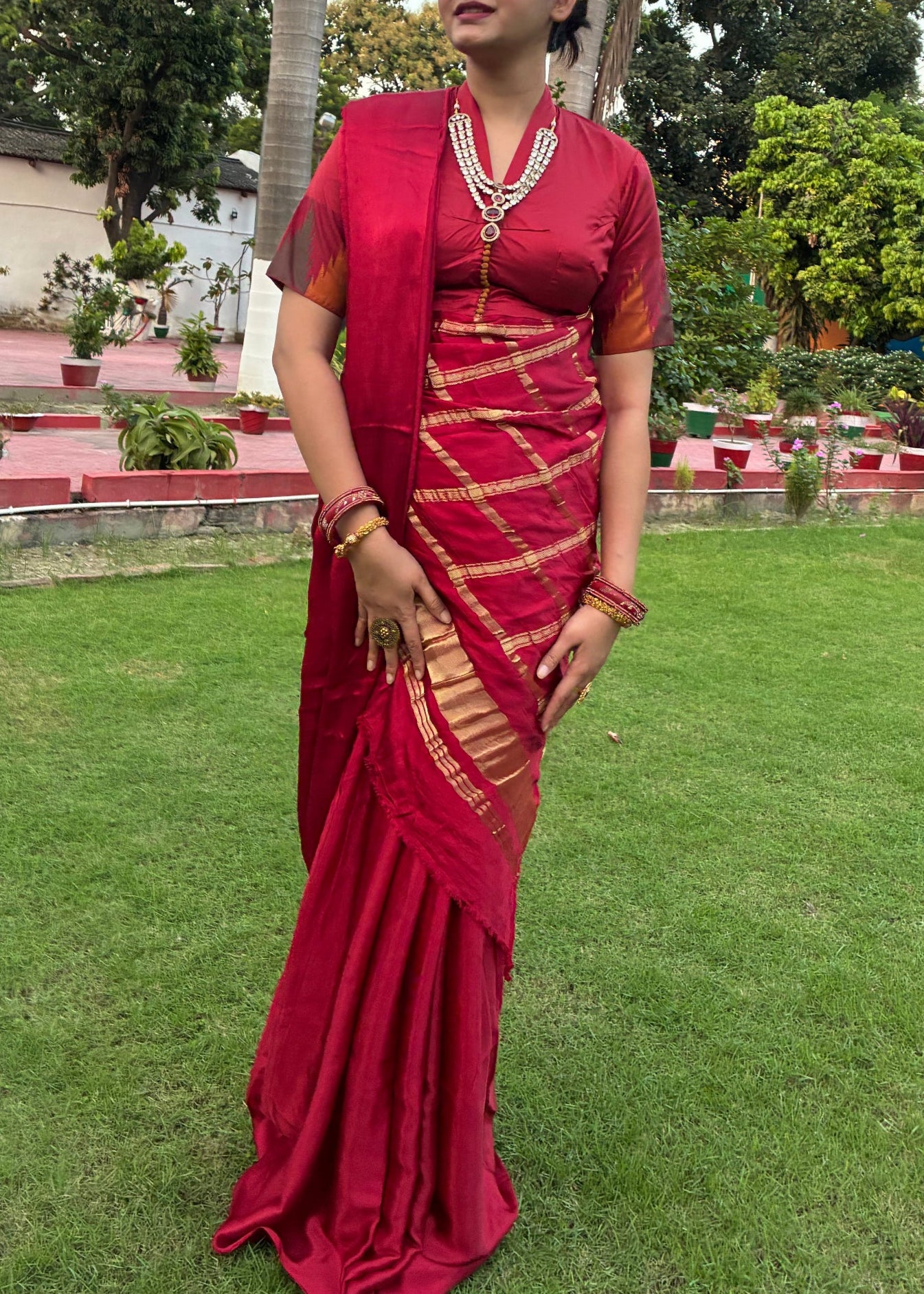 Woman in a red saree standing in a park with green grass and trees.