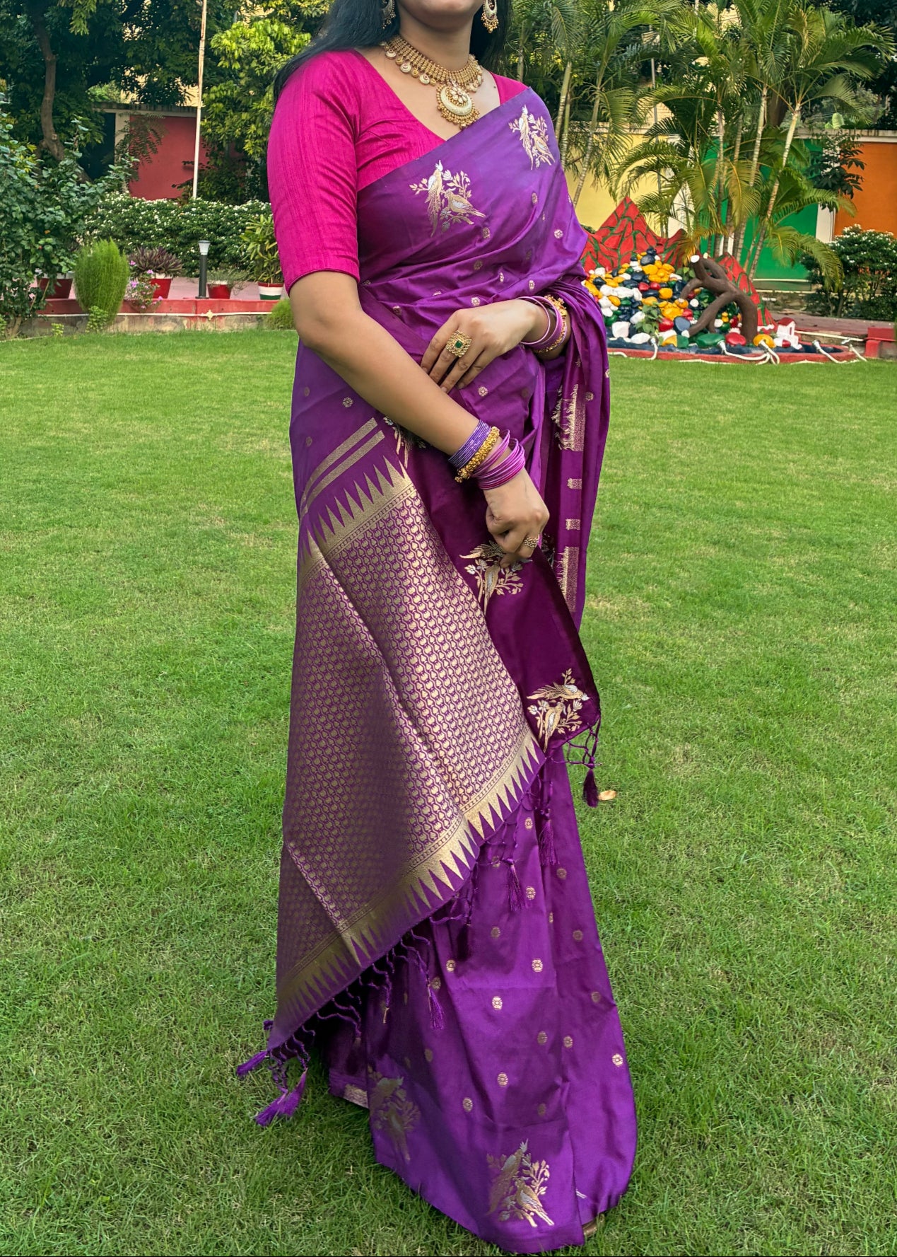 Woman in a purple saree standing on grass with trees in the background