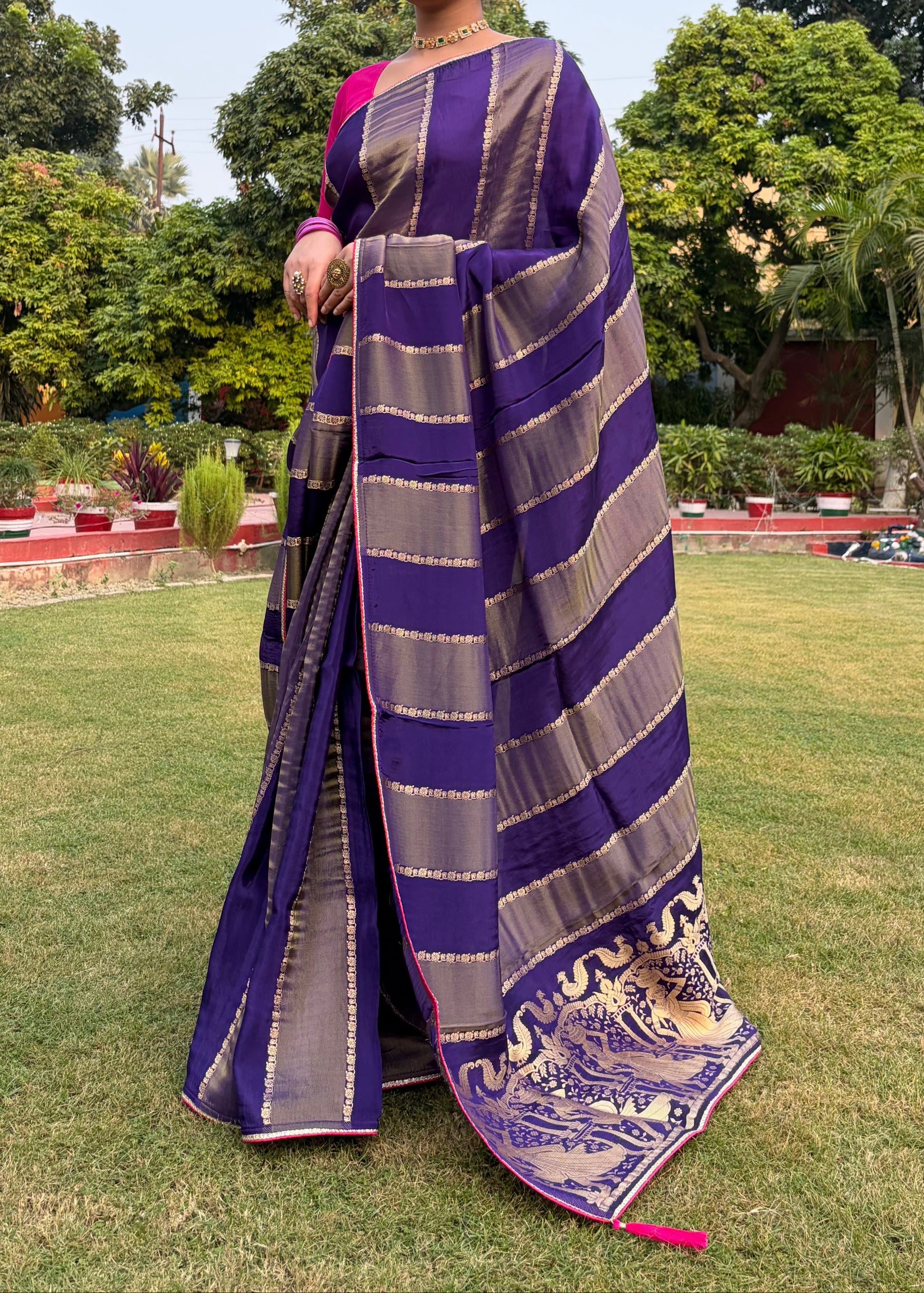 Woman wearing a purple and gray striped saree in an outdoor setting with greenery.