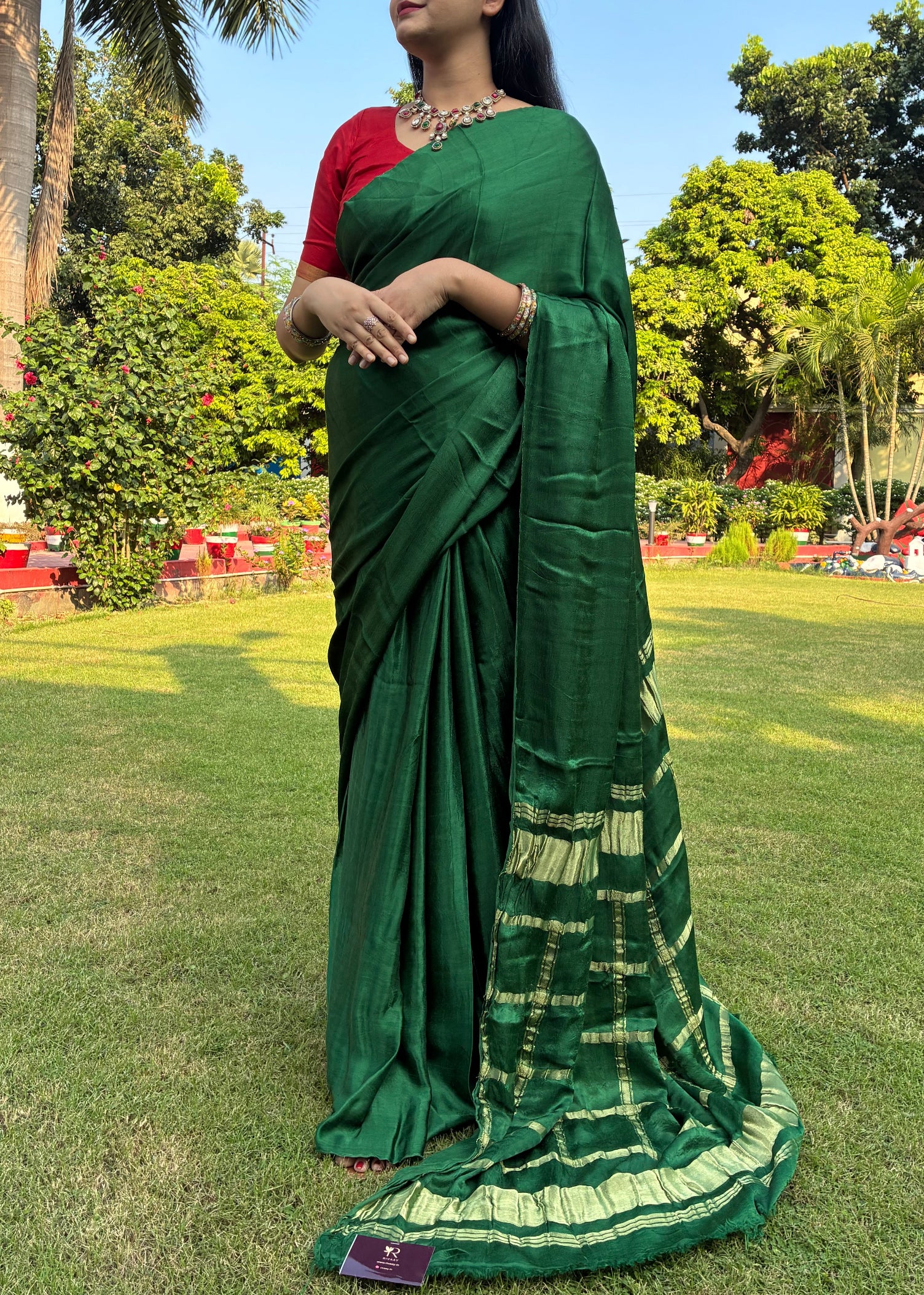 Woman in a green saree standing on grass with trees and people in the background