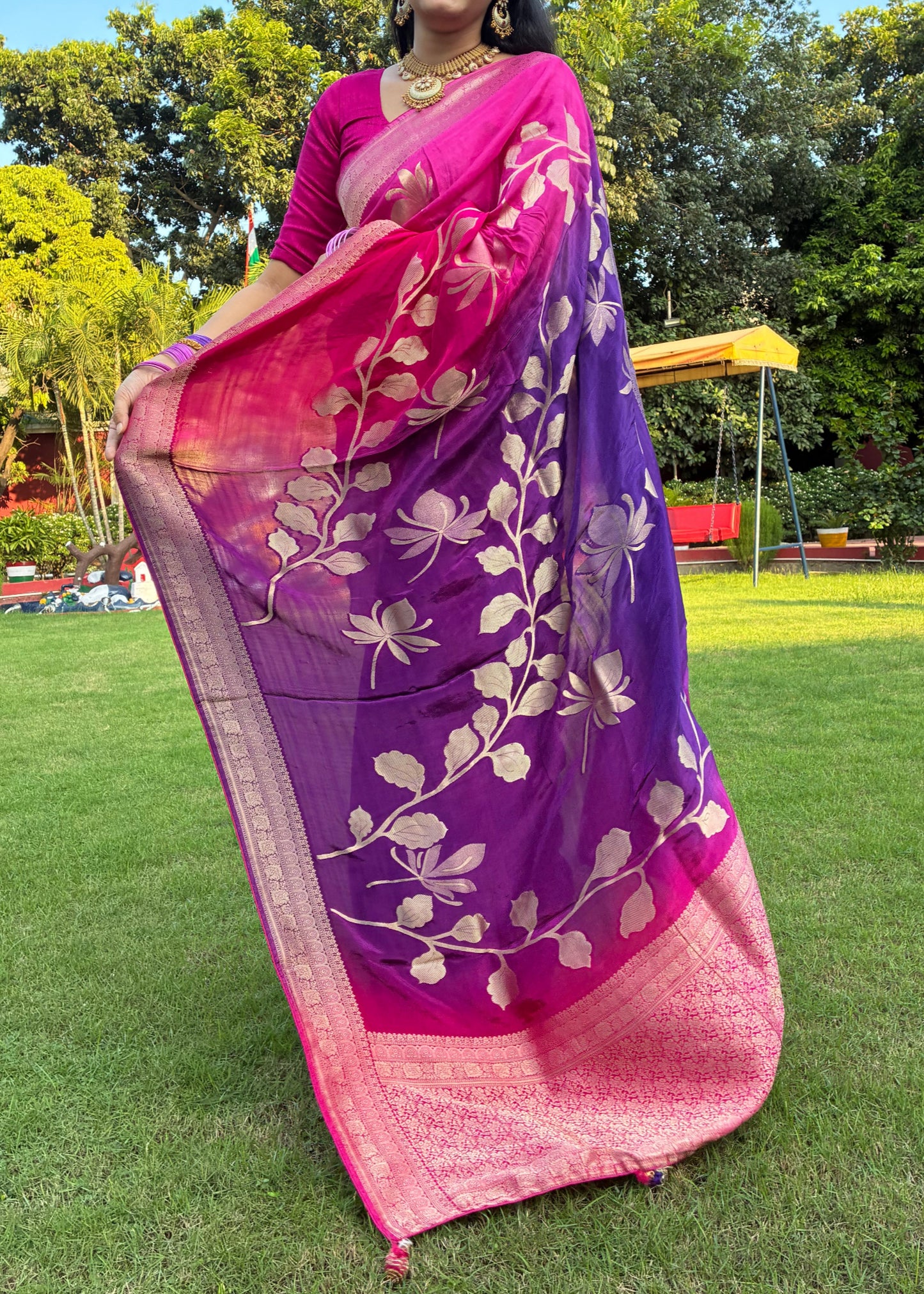 Woman in a pink and purple saree with floral patterns standing outdoors on a grassy area with trees in the background.