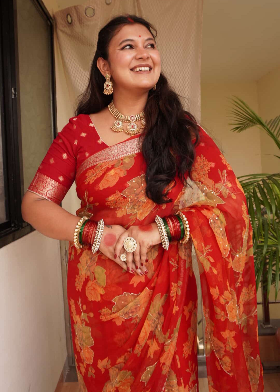 Woman in a red saree with gold patterns standing indoors.